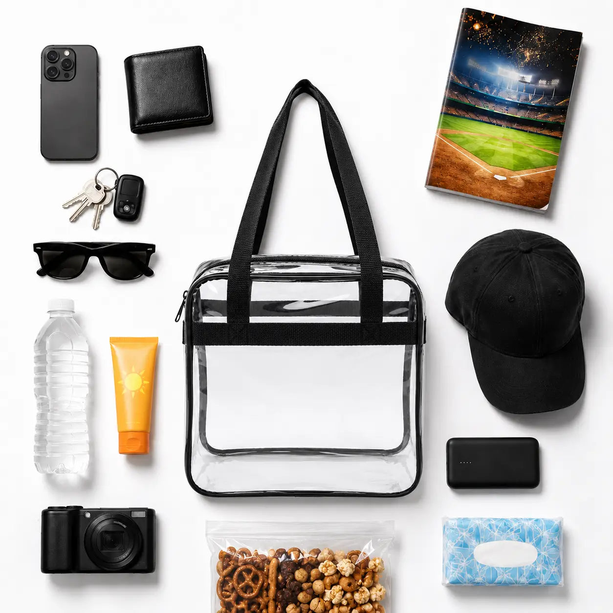 A clear tote bag with black trim and handles is arranged in a flat lay with various everyday items around it on a white background, including a smartphone, wallet, keys, sunglasses, water bottle, sunscreen, camera, snack bag, tissue, and a baseball stadium notebook.