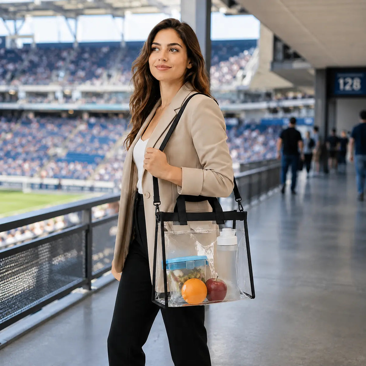 A woman in a beige blazer and black pants carries a clear tote bag with black trim and handles, filled with snacks, while walking through a stadium corridor.
