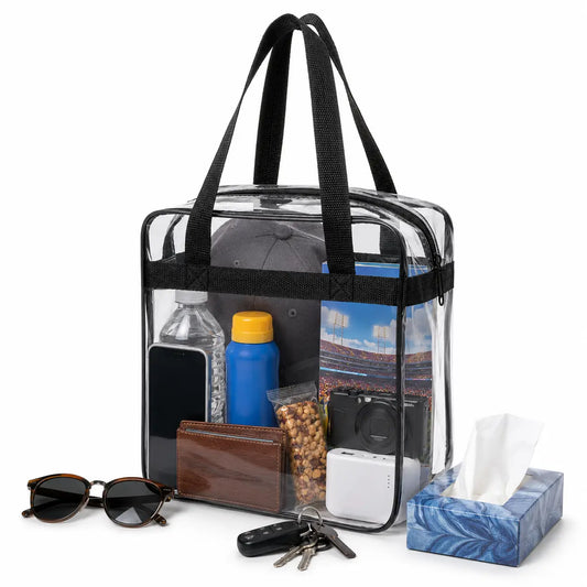 A clear plastic carry bag with black handles and trim, shown filled with assorted items including a phone, water bottle, wallet, keys, sunglasses, tissue box, and camera. The bag is photographed on a clean white background.