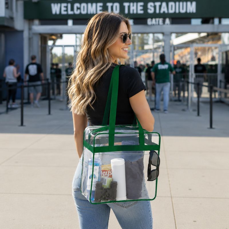 Woman carrying the clear stadium tote with green trim over her shoulder while walking toward a stadium entrance gate.