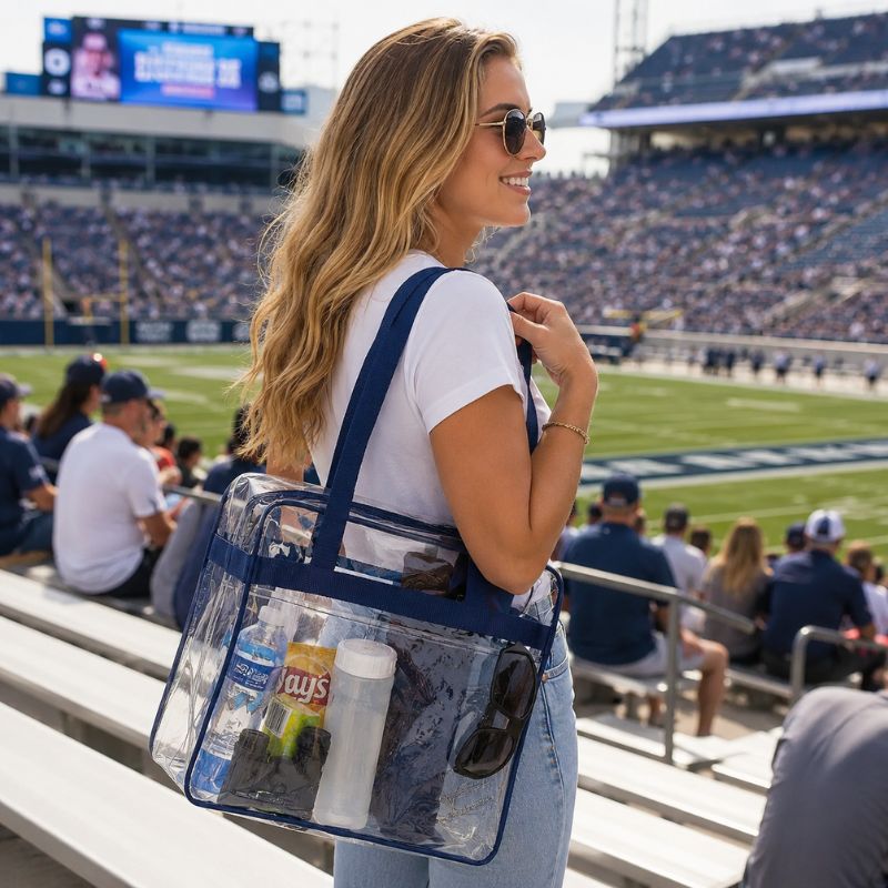 Woman carrying the clear stadium tote with navy trim over her shoulder while seated on stadium bleachers.